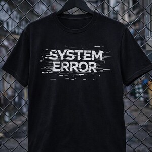 Może przedstawiać: Czarny t-shirt z białym napisem „SYSTEM ERROR” z efektem glitch. Koszulka wisi na czarnym wieszaku na tle ogrodzenia z siatki drucianej.