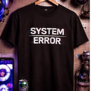 Könnte beinhalten: Schwarzes T-Shirt mit dem weißen Schriftzug "SYSTEM ERROR" mit Glitch-Effekt. Das Shirt hängt an einem Holzbügel. Gaming-Zubehör und eine Getränkedose sind im Hintergrund zu sehen.