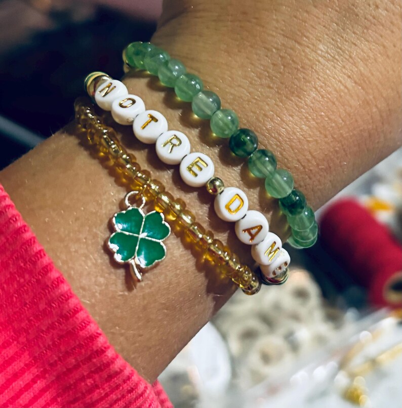 K&ouml;nnte beinhalten: Drei Armb&auml;nder, die an einem Handgelenk &uuml;bereinander liegen. Das obere Armband ist gr&uuml;n und besteht aus Perlen. Das mittlere Armband ist golden und besteht aus Perlen. Das untere Armband ist wei&szlig; mit goldenen Buchstaben, die "NOTRE DAME" buchstabieren, und hat einen gr&uuml;nen Gl&uuml;cksbringer mit vier Bl&auml;ttern.