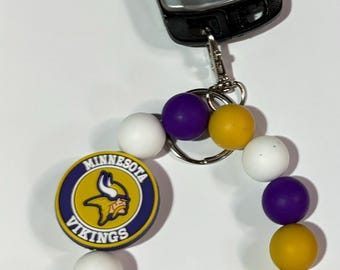 Puede incluir: Un llavero de cuentas moradas, blancas y doradas con un dije con el logotipo de los Minnesota Vikings. El llavero está unido a un llavero de coche plateado.