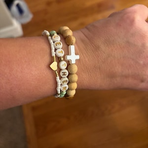 Può includere: Un set di tre braccialetti con perline di legno, perline bianche e un piccolo ciondolo a forma di cuore. I braccialetti compongono la scritta "Let Them" e presentano un ciondolo a forma di croce bianca.