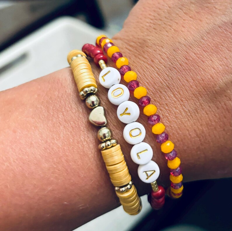 K&ouml;nnte beinhalten: Ein Set aus drei Armb&auml;ndern mit einem goldenen Herz-Anh&auml;nger, einem mehrlagigen Perlenarmband mit gelben und burgunderfarbenen Perlen und einem wei&szlig;en Perlenarmband mit dem in goldenen Buchstaben geschriebenen Wort "LOYOLA".