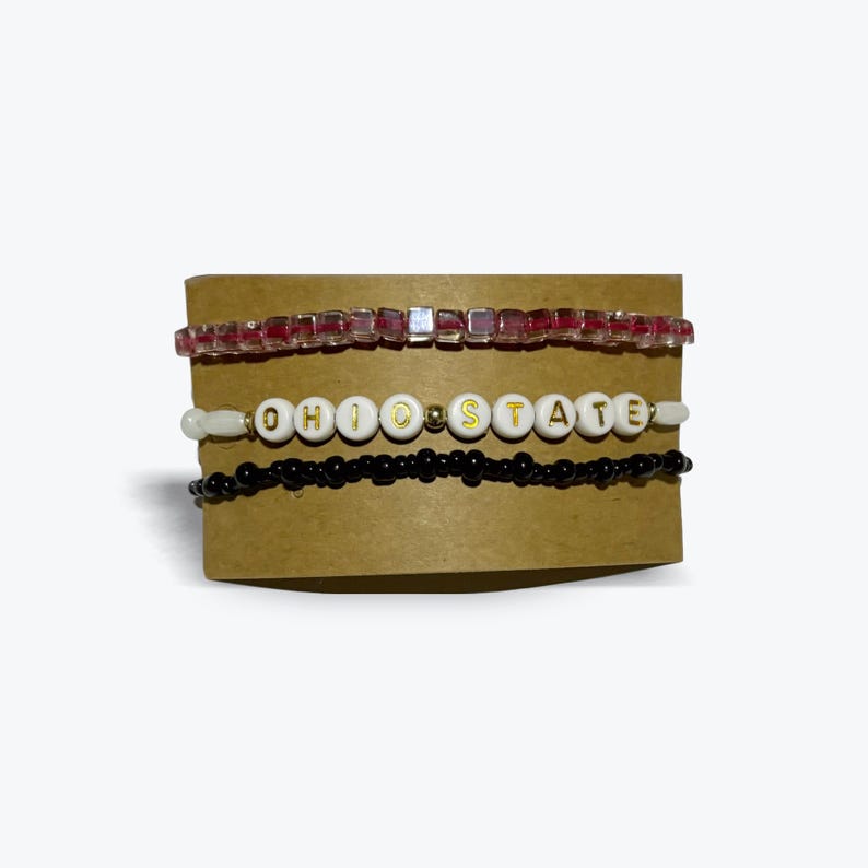 K&ouml;nnte beinhalten: Drei Perlenarmb&auml;nder, eines mit roten und klaren Perlen, eines mit schwarzen Perlen und eines mit wei&szlig;en Perlen, die "OHIO STATE" buchstabieren.