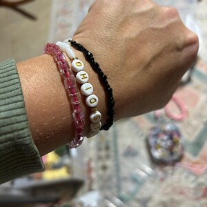 Peut inclure: Un poignet avec quatre bracelets. Le bracelet du haut est noir avec de petites perles noires. Le deuxième bracelet est blanc avec le mot "OHIO" écrit en lettres dorées. Le troisième bracelet est rose avec de petites perles roses. Le bracelet du bas est transparent avec de petites perles transparentes.
