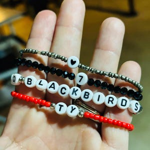 Könnte beinhalten: Vier Armbänder mit Perlen in Silber, Schwarz und Rot. Ein Armband schreibt "BLACKBIRDS" mit weißen Buchstabenperlen. Andere Armbänder zeigen ein Herz, die Zahl 7 und andere Buchstabenperlen.