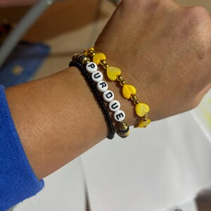 K&ouml;nnte beinhalten: Drei Armb&auml;nder, die auf einem Handgelenk &uuml;bereinander liegen. Das untere Armband ist schwarz mit kleinen schwarzen Perlen und wei&szlig;en Buchstabenperlen, die "PURDUE" buchstabieren. Das mittlere Armband ist golden mit gelben herzf&ouml;rmigen Perlen. Das obere Armband ist golden mit gelben herzf&ouml;rmigen Perlen.