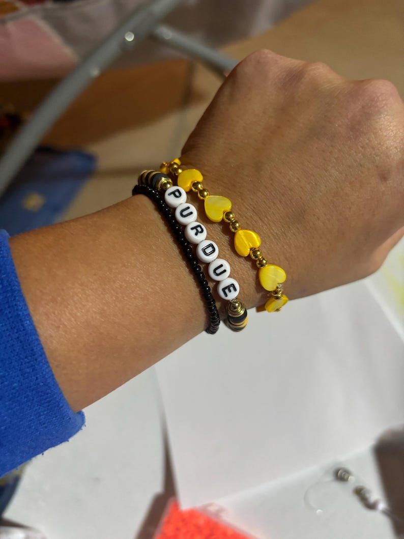 K&ouml;nnte beinhalten: Drei Armb&auml;nder, die auf einem Handgelenk &uuml;bereinander liegen. Das untere Armband ist schwarz mit kleinen schwarzen Perlen und wei&szlig;en Buchstabenperlen, die "PURDUE" buchstabieren. Das mittlere Armband ist golden mit gelben herzf&ouml;rmigen Perlen. Das obere Armband ist golden mit gelben herzf&ouml;rmigen Perlen.