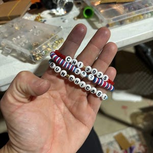 Könnte beinhalten: Drei Perlenarmbänder mit den Worten "CHI 2025", "RUN" und "CHICAGO" in weißen Buchstaben auf blauem und rotem Hintergrund.