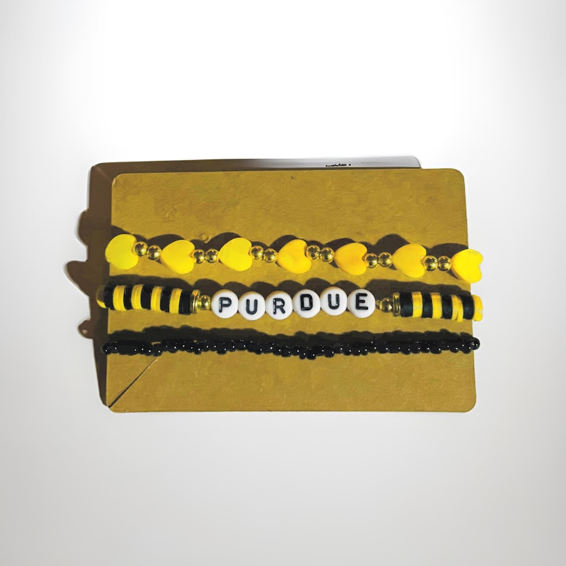 K&ouml;nnte beinhalten: Drei Perlenarmb&auml;nder mit einem schwarz-gelben Streifenmuster, ein schwarzes Perlenarmband mit dem Wort "PURDUE" in wei&szlig;en Buchstaben und ein gelbes Perlenarmband in Herzform.