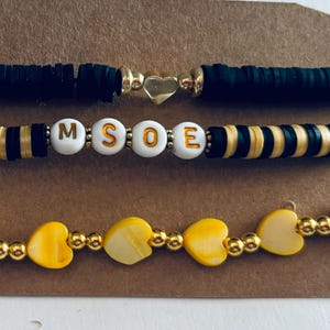 K&ouml;nnte beinhalten: Drei Armb&auml;nder mit schwarzen und goldenen Perlen. Das obere Armband ist schwarz mit einem goldenen Herz-Anh&auml;nger. Das mittlere Armband hat schwarz-goldene gestreifte Perlen mit wei&szlig;en Buchstabenperlen, die "MSOE" buchstabieren. Das untere Armband hat gelbe Herzperlen mit goldenen Perlen.