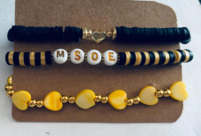 K&ouml;nnte beinhalten: Drei Armb&auml;nder mit schwarzen und goldenen Perlen. Das obere Armband ist schwarz mit einem goldenen Herz-Anh&auml;nger. Das mittlere Armband hat schwarz-goldene gestreifte Perlen mit wei&szlig;en Buchstabenperlen, die "MSOE" buchstabieren. Das untere Armband hat gelbe Herzperlen mit goldenen Perlen.