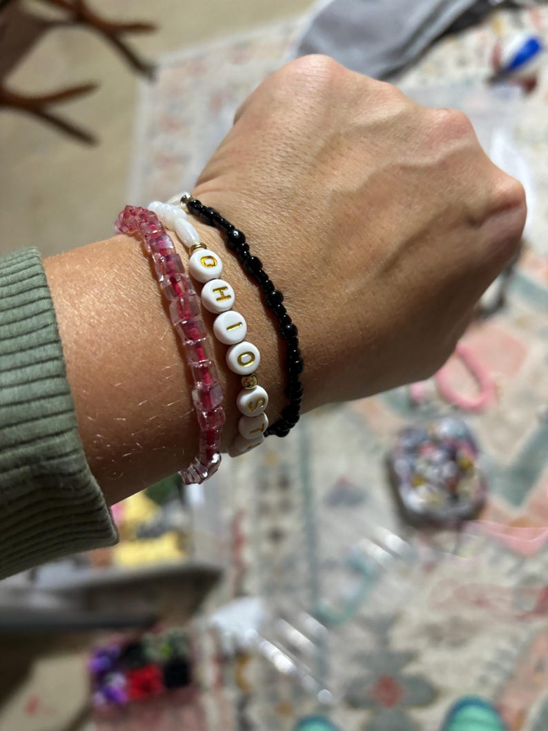 K&ouml;nnte beinhalten: Ein Handgelenk mit vier Armb&auml;ndern. Das oberste Armband ist schwarz mit kleinen schwarzen Perlen. Das zweite Armband ist wei&szlig; mit dem Wort "OHIO" in goldenen Buchstaben. Das dritte Armband ist rosa mit kleinen rosa Perlen. Das unterste Armband ist klar mit kleinen klaren Perlen.