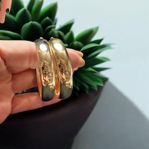 May include: A pair of gold-colored hoop earrings. The earrings are held in front of a green plant. The earrings have a smooth, polished surface and a rounded shape. The earrings are a classic jewelry item.