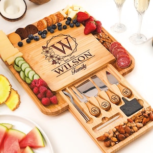 May include: A square bamboo charcuterie board featuring a slide-out drawer containing three metal cheese knives and a fork with wooden handles engraved with the letter W. The board is laser-engraved with a floral design, the letter W, and the text WILSON Family EST. 2025.