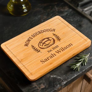 May include: A rectangular wooden cutting board with the words "MOM'S SOURDOUGH" and "Sarah Wilson" engraved on it. The board features a bread illustration and the year "Est. 2023". The board is light brown and has a juice groove.