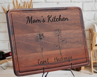 May include: A rectangular wooden cutting board with a dark brown finish. The words "Mom's Kitchen" are engraved at the top, with floral illustrations and names below. The board is displayed on a black metal stand.