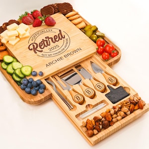 May include: A wooden charcuterie board with a slide-out drawer containing cheese knives and a fork. The board is filled with snacks, including cheese, strawberries, blueberries, and cookies. The board is engraved with the words "Officially Retired 2025".