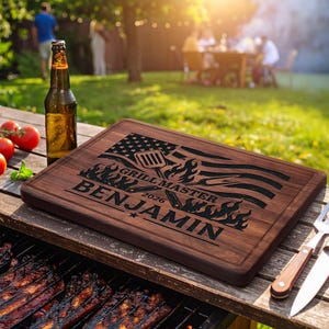 May include: A rectangular wooden cutting board with a juice groove, engraved with a stylized American flag, flames, a spatula, and the text "GRILL MASTER 2026 BENJAMIN". The board sits on a grill next to a brown glass beer bottle and tomatoes.