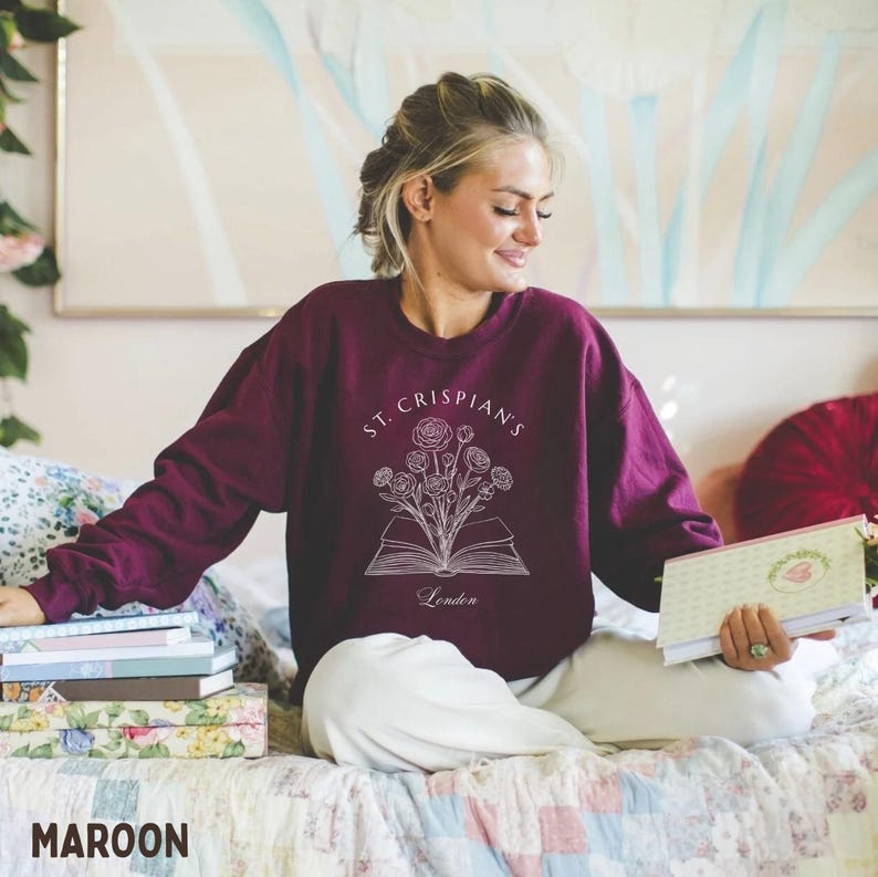 May include: A maroon long-sleeve crewneck sweatshirt featuring a white line art graphic of an open book with flowers emerging from the pages. The text "ST. CRISPIAN'S" is printed in an arc above the illustration, with "London" printed below.