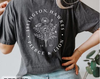 Può includere: Una t-shirt a maniche corte grigio antracite con una grafica a linee bianche raffigurante un bouquet di fiori racchiuso in una cornice ovale. Il testo "ISLINGTON, HAWKES" e "PIERCE & LION" è stampato attorno all'ovale con i numeri "18" e "83".