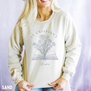 May include: A sand-coloured crewneck sweatshirt featuring a dark grey line art graphic of an open book with a bouquet of flowers emerging from the pages. The text "ST. CRISPIAN'S" is arched above the illustration, with the word "London" below.