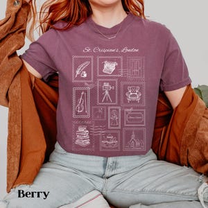 May include: Berry-coloured t-shirt with a stamp-like design featuring various illustrations and the text "St. Crispian's, London." The design includes images of a feather, typewriter, door, violin, film camera, armchair, books, and a church.