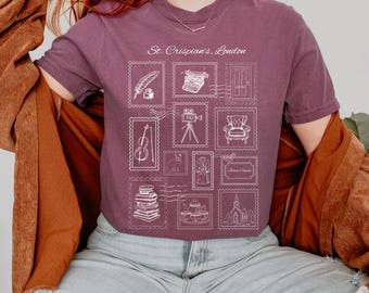 Può includere: T-shirt color bacca con un design a forma di timbro con varie illustrazioni e la scritta "St. Crispian's, London". Il design include immagini di una piuma, macchina da scrivere, porta, violino, cinepresa, poltrona, libri e una chiesa.