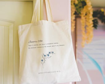 May include: Cream-colored tote bag with long handles, hanging on a door. The bag features the text "January 24th" and a floral illustration in blue. Additional text is printed below the illustration.
