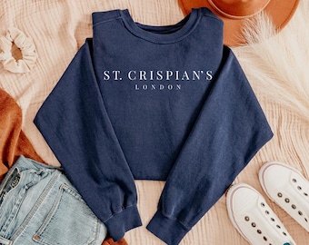 Könnte beinhalten: Ein marineblaues Sweatshirt mit dem weißen Text "ST. CRISPIAN'S LONDON". Das Sweatshirt wird mit einer Jeans, weißen Turnschuhen und einem braunen Hut präsentiert. Der Hintergrund ist ein hellbrauner Stoff.