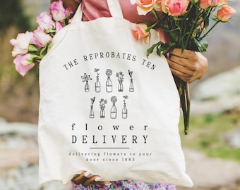 May include: A white canvas tote bag featuring black line art of flowers in bottles and the text THE REPROBATES TEN flower DELIVERY delivering flowers to your door since 1883. The bag is held against a pink shirt and a bouquet of pink roses.