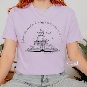 May include: Lilac t-shirt featuring a black illustration of a ship emerging from an open book, with stars and a crescent moon. The text reads "But some day you will be old enough to start reading fairy tales again" and "C.S. Lewis".