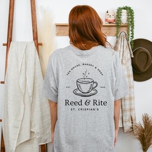 Pu&ograve; includere: Una maglietta grigio chiaro con una grafica nera di una tazza da t&egrave; e un piattino, con il testo "TEA HOUSE, BAKERY & SHOP" e "Reed & Rite ST. CRISPIAN'S". La maglietta &egrave; indossata da una persona.