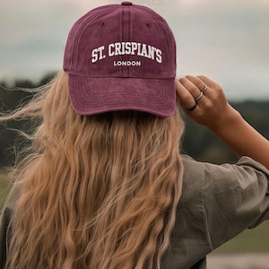 May include: A maroon baseball cap with the text "ST. CRISPIAN'S LONDON" in white lettering. The cap is worn by a person with long, wavy blonde hair. The person is wearing a green shirt.