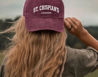May include: A maroon baseball cap with the text "ST. CRISPIAN'S LONDON" in white lettering. The cap is worn by a person with long, wavy blonde hair. The person is wearing a green shirt.