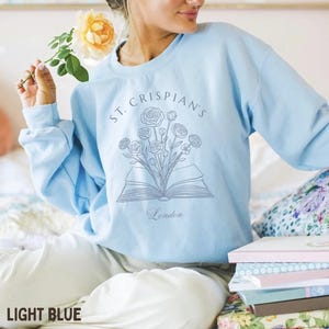 May include: A light blue crewneck sweatshirt featuring a dark blue line art graphic of an open book with a bouquet of flowers emerging from the pages. The text "ST. CRISPIAN'S" is arched above the illustration, with the word "London" below.