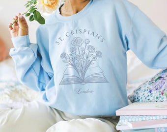 Könnte beinhalten: Ein hellblaues Sweatshirt mit Rundhalsausschnitt und einer dunkelblauen Strichzeichnung eines aufgeschlagenen Buches, aus dessen Seiten ein Blumenstrauß wächst. Der Text "ST. CRISPIAN'S" ist über der Illustration gebogen, darunter steht "London".
