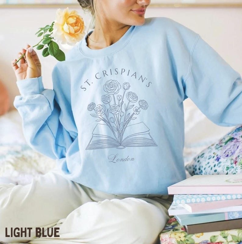 May include: A light blue crewneck sweatshirt featuring a dark blue line art graphic of an open book with a bouquet of flowers emerging from the pages. The text "ST. CRISPIAN'S" is arched above the illustration, with the word "London" below.