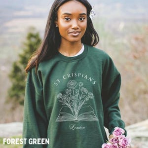 May include: A forest green crewneck sweatshirt featuring a white line art graphic of an open book with a bouquet of flowers emerging from the pages. The text "ST. CRISPIAN'S" is arched above the illustration, and the word "London" is printed below.