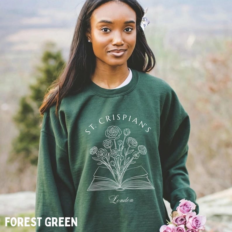 May include: A forest green crewneck sweatshirt featuring a white line art graphic of an open book with a bouquet of flowers emerging from the pages. The text "ST. CRISPIAN'S" is arched above the illustration, and the word "London" is printed below.