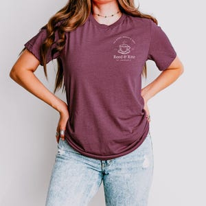 Pu&ograve; includere: T-shirt bordeaux a maniche corte con un piccolo logo bianco raffigurante una tazza da t&egrave; e un piattino, e la scritta "Reed & Rite". La modella indossa jeans azzurri.