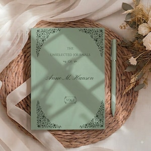 Op de afbeelding: Een saliegroen notitieboek met harde kaft, donkergroene florale hoekaccenten en de tekst "THE UNSELECTED JOURNALS OF Anne M. Hansen VOL I" op de voorkant, vergezeld van een bijpassende saliegroene pen die verticaal aan de rechterkant is geplaatst.