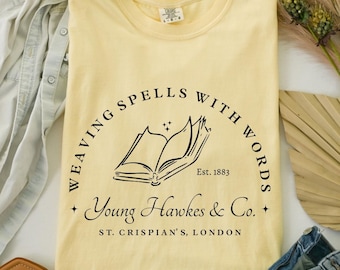 May include: A light yellow t-shirt with a black graphic of an open book and the text "WEAVING SPELLS WITH WORDS" and "Young Hawkes & Co.". The shirt also has the text "ST. CRISPIAN'S, LONDON" and "Est. 1883".