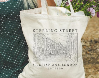 Puede incluir: Bolso tote de color crema con el texto "STERLING STREET" sobre una ilustración en blanco y negro de una escena callejera. Debajo de la ilustración está "ST. CRISPIAN'S, LONDON EST 1800". El bolso es llevado por una persona y contiene flores.