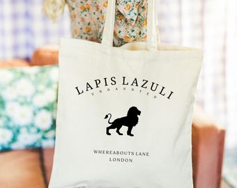Puede incluir: Bolso tote de color crema con las palabras "LAPIS LAZULI UNDAUNTED" en una fuente curva sobre una silueta de león negro. Debajo del león está "WHEREABOUTS LANE LONDON". El bolso tiene dos asas largas.