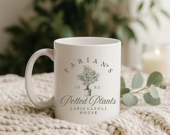 May include: White ceramic mug with a curved handle. The mug features a green illustration of a potted plant and the text "PARIAN'S 1883 Potted Plants LAPIS LAZULI HOUSE". The mug is on a cream-coloured knitted blanket.
