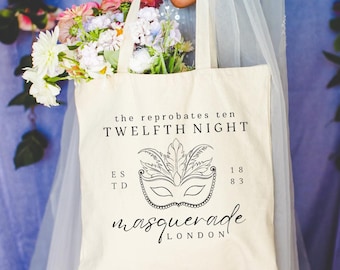 Puede incluir: Bolso tote color crema con texto negro y un diseño de máscara de carnaval. El texto dice "the reprobates ten TWELFTH NIGHT" y "masquerade LONDON". La bolsa está llena de un ramo de flores coloridas.