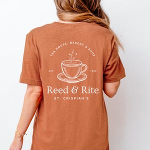 Pu&ograve; includere: Una maglietta color ruggine con una grafica bianca sul retro. La grafica include una tazza da t&egrave; e un piattino, le parole "TEA HOUSE, BAKERY & SHOP" e il testo "Reed & Rite". La maglietta ha anche il testo "ST. CRISPIAN'S".