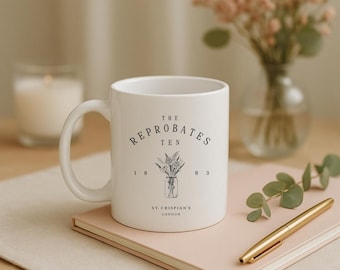 May include: White ceramic mug with the text "THE REPROBATES TEN 1883 ST. CRISPIAN'S LONDON" and a floral illustration. The mug is on a pink notebook with a gold pen, with a candle and flowers in the background.