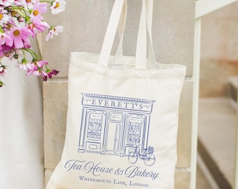 Puede incluir: Bolso tote de color crema con una ilustración azul de una fachada de tienda, una bicicleta y el texto "Everett's Tea House & Bakery, Whereabouts Lane, London." El bolso tiene dos asas y está siendo sostenido.
