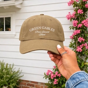 May include: A khaki-coloured baseball cap with the text "Green Gables Prince Edward Island" in white. The cap is held up by a hand with a ring. The background includes a white house and pink flowers.